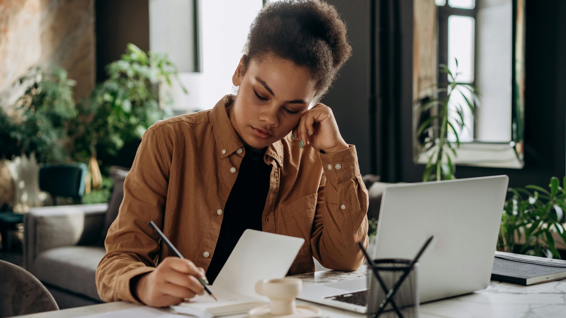 A dedicated woman with curly hair writing in a notebook at a modern office desk.