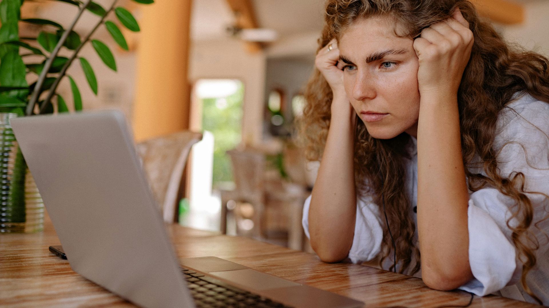 A woman with curly hair intensely focusing on her laptop indoors with plants and natural lighting.