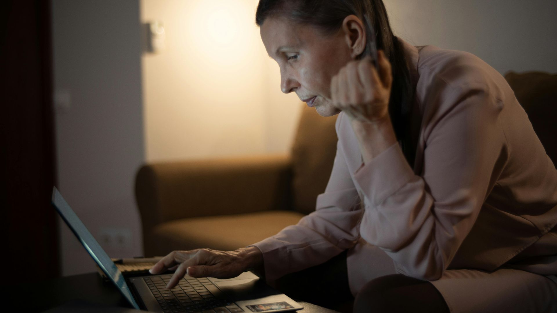 Senior woman using laptop in a dimly lit living room, focused on screen.