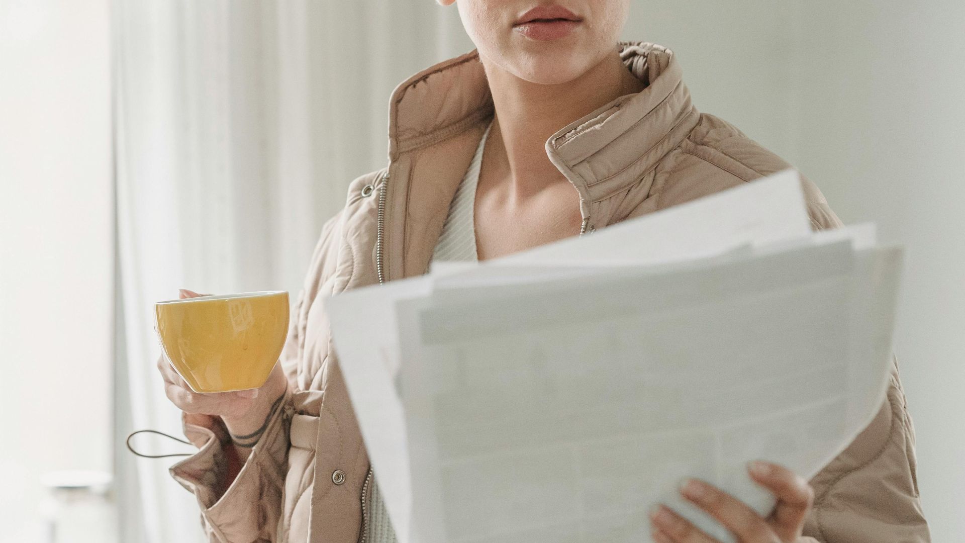 A woman stands by a window holding papers and a coffee cup, in daylight.