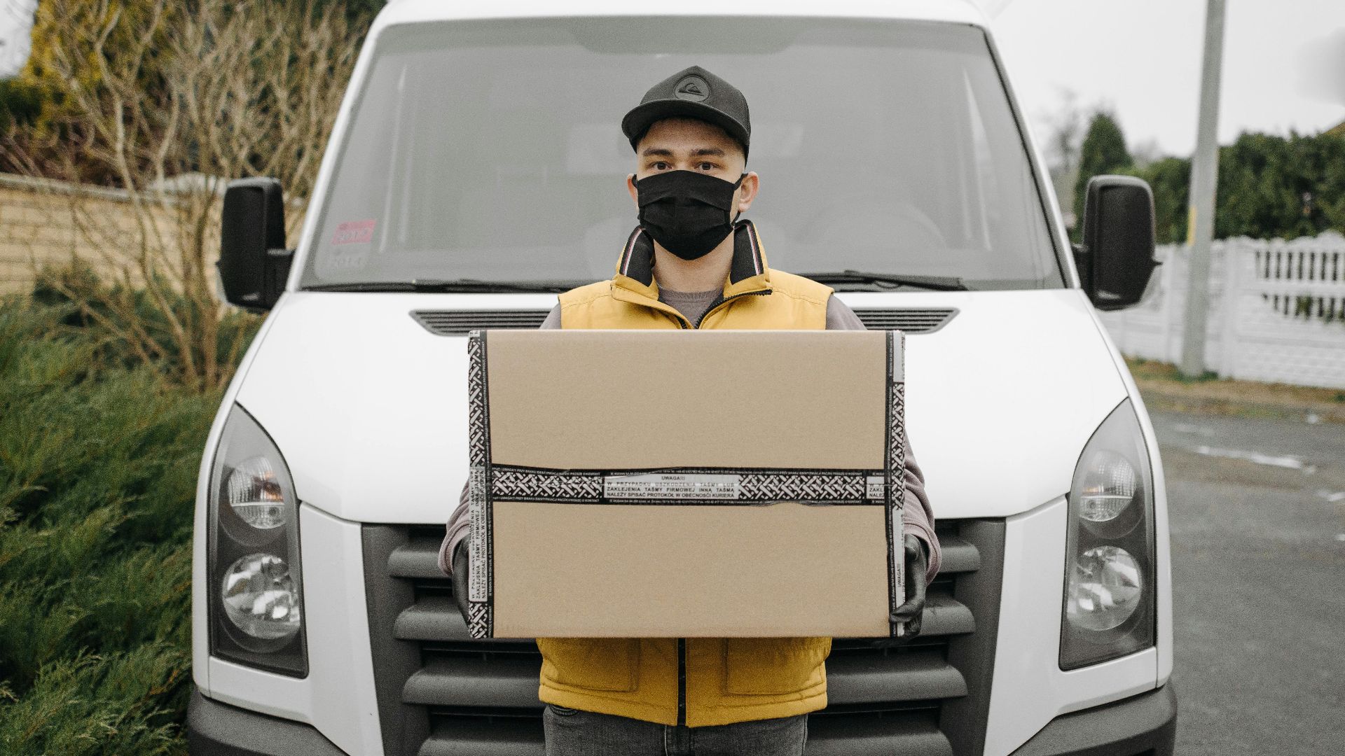 A deliveryman wearing a face mask holding a package in front of a van outdoors.