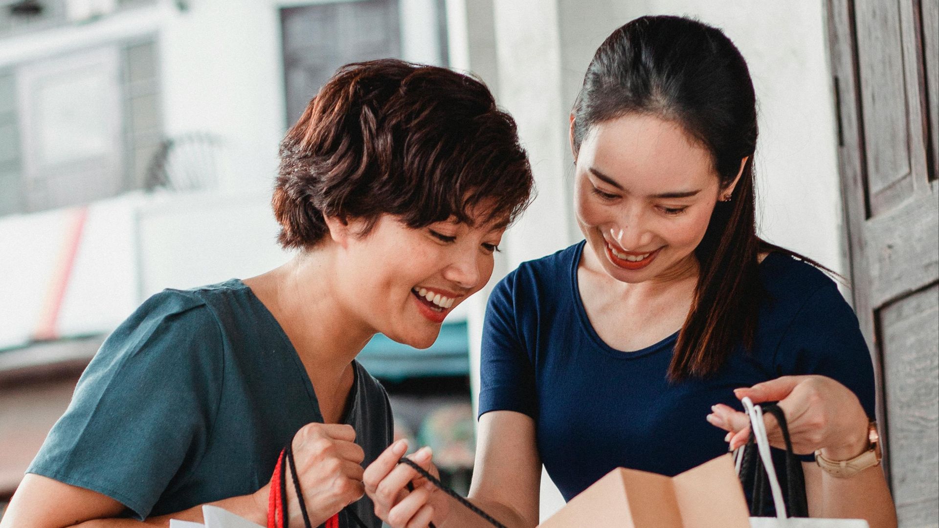 Ethnic female friends in casual clothes holding shopping bags and looking at goods while standing on street and smiling