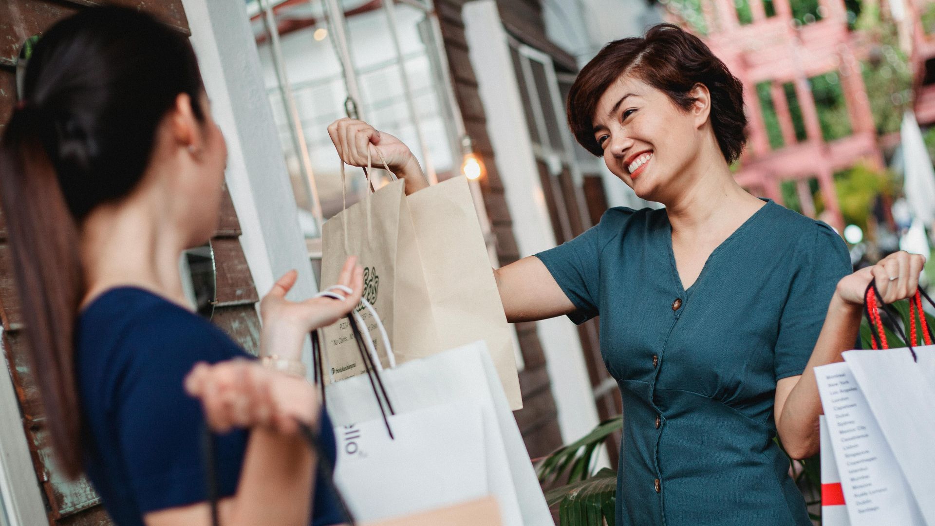 Two women smiling and holding shopping bags outdoors, enjoying a sunny day.