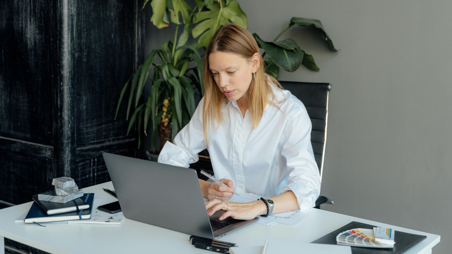 Focused woman in white shirt working on laptop at desk with plants and books.