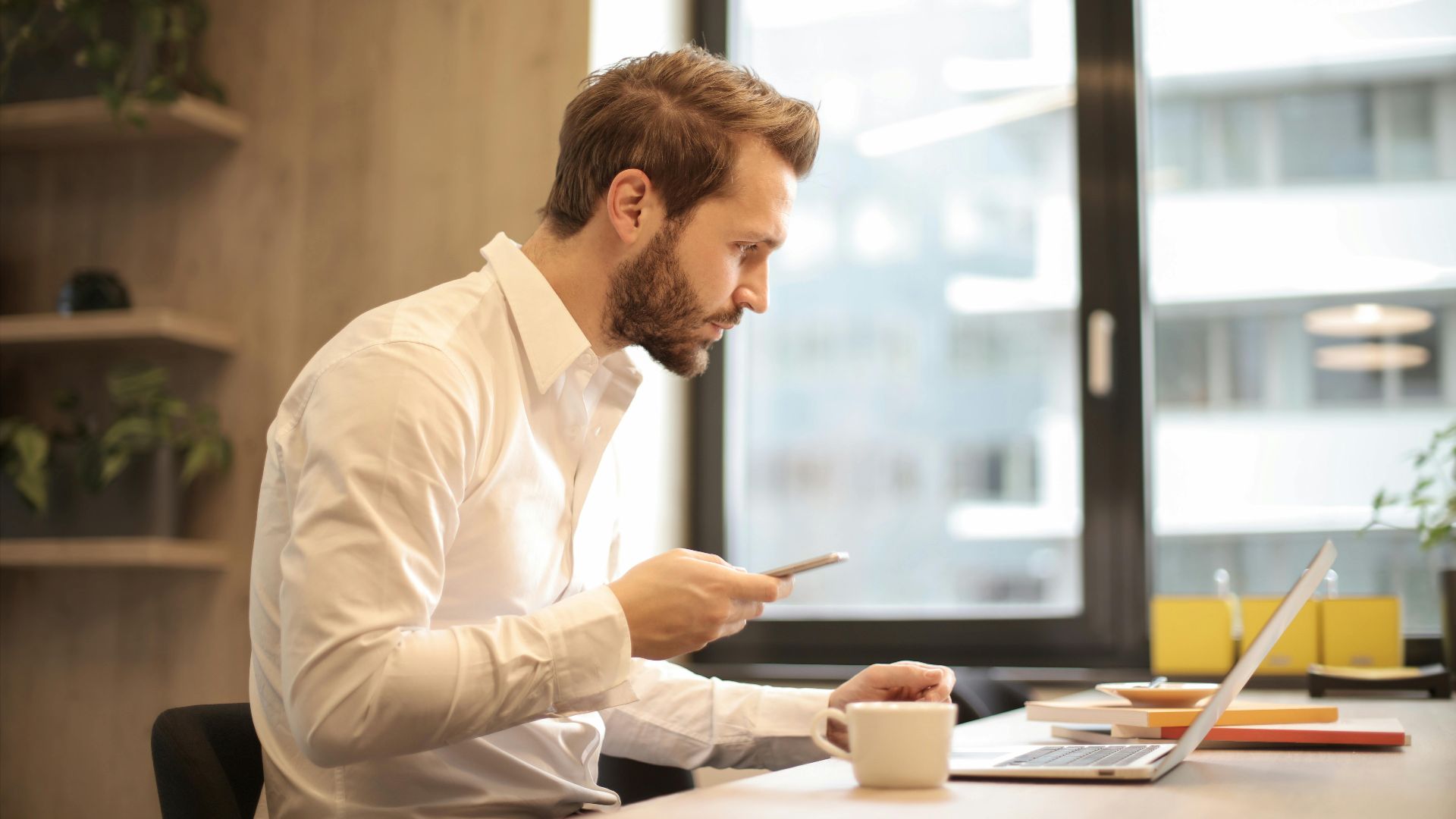 Focused businessman working on laptop while checking smartphone in modern office.