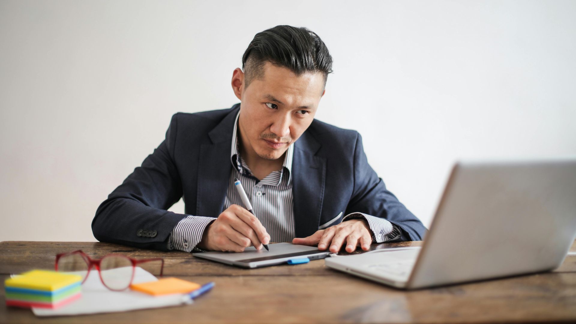 Concentrated Asian man using a digital tablet at his desk, focusing on work in a modern office setting.
