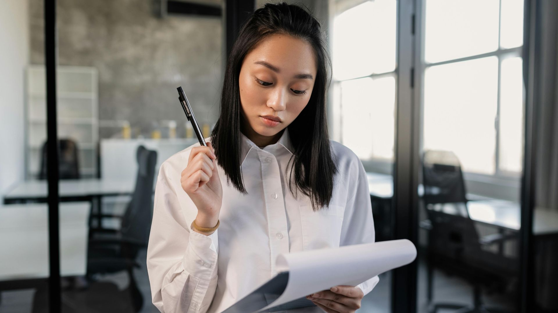 Confident Asian woman in office reviewing documents with pen.