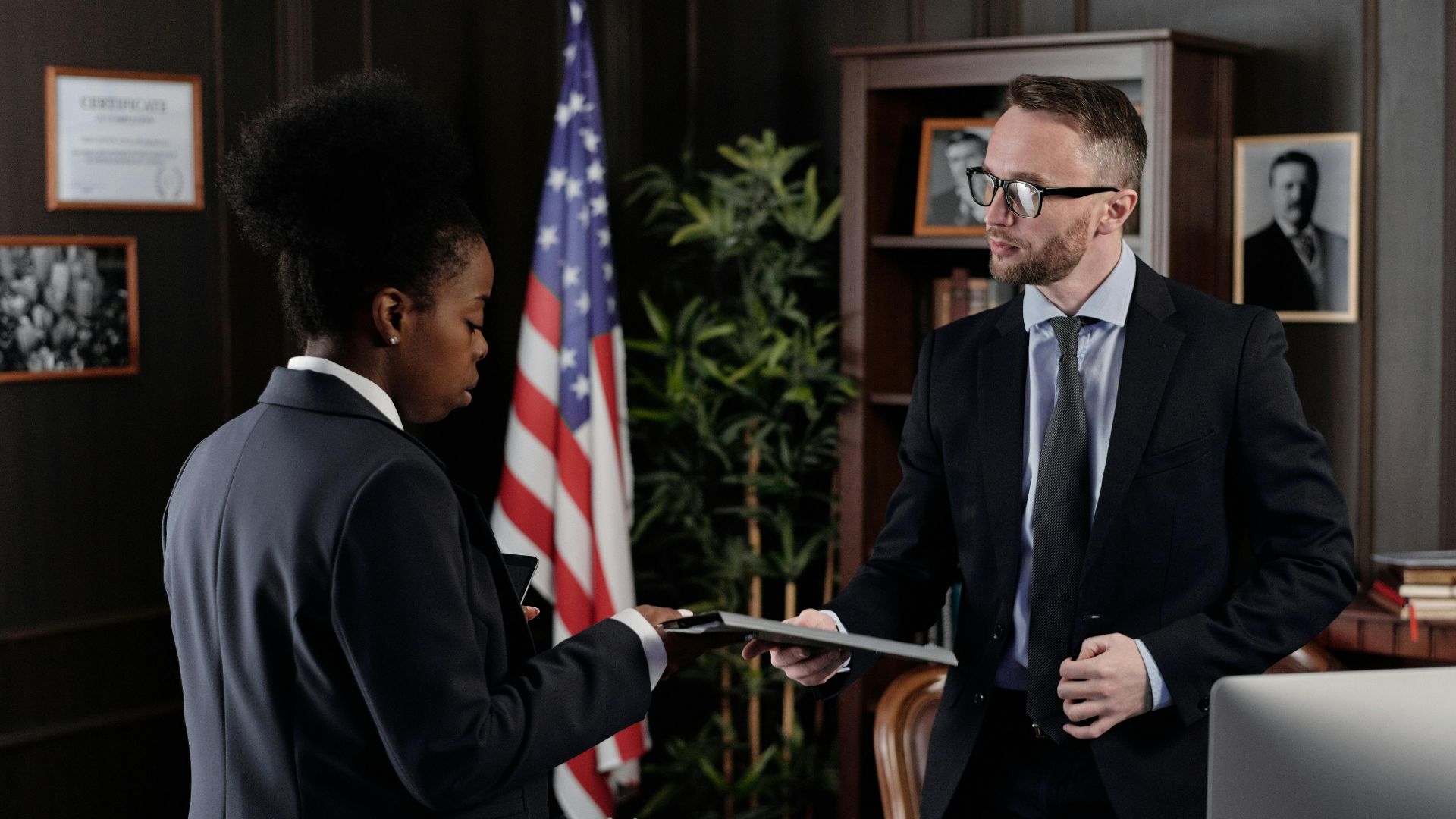 Business professionals engage in a meeting in a formal office environment with an American flag in the background.