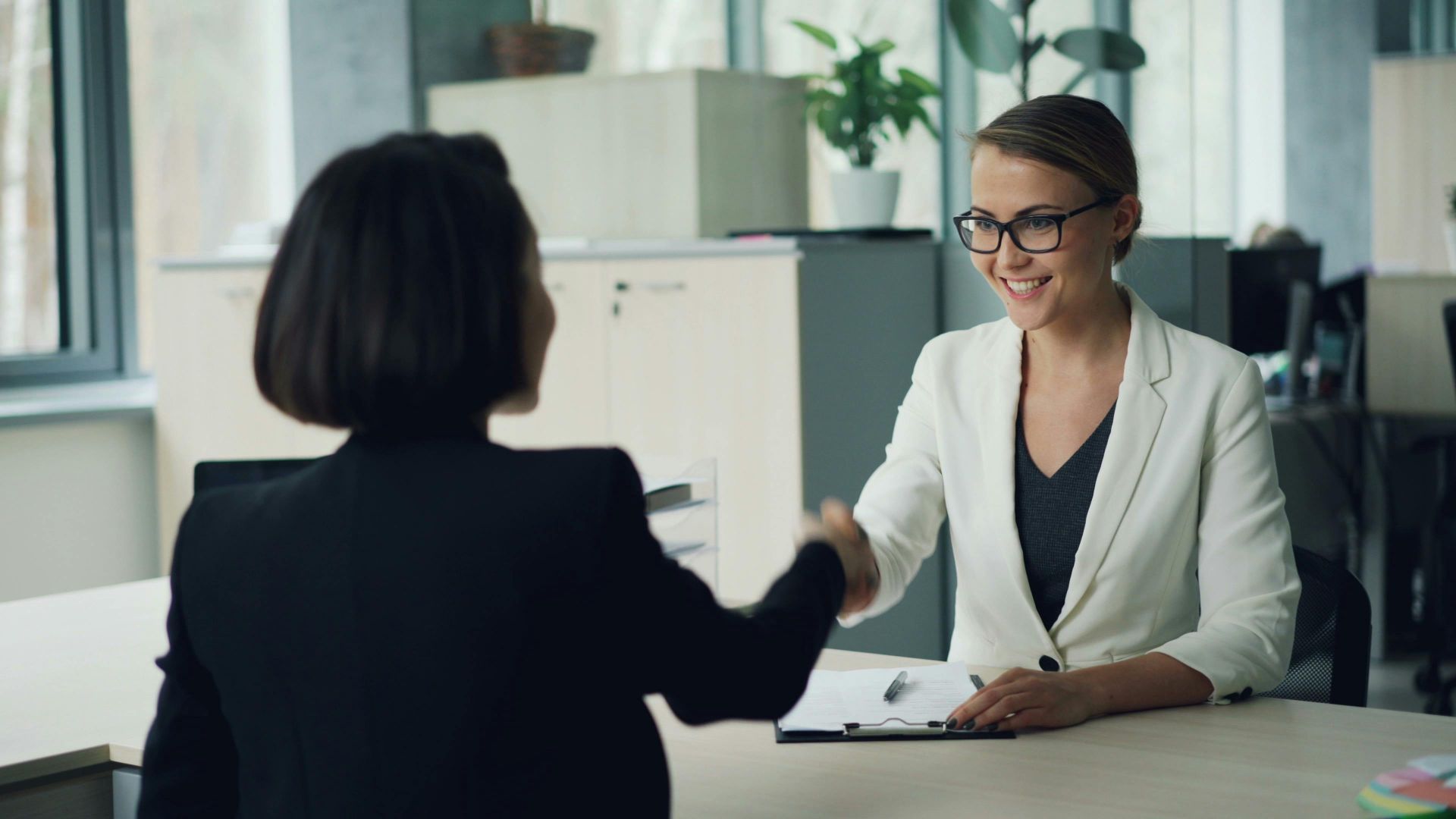 Two businesswomen shaking hands in a modern office setting, symbolizing partnership.
