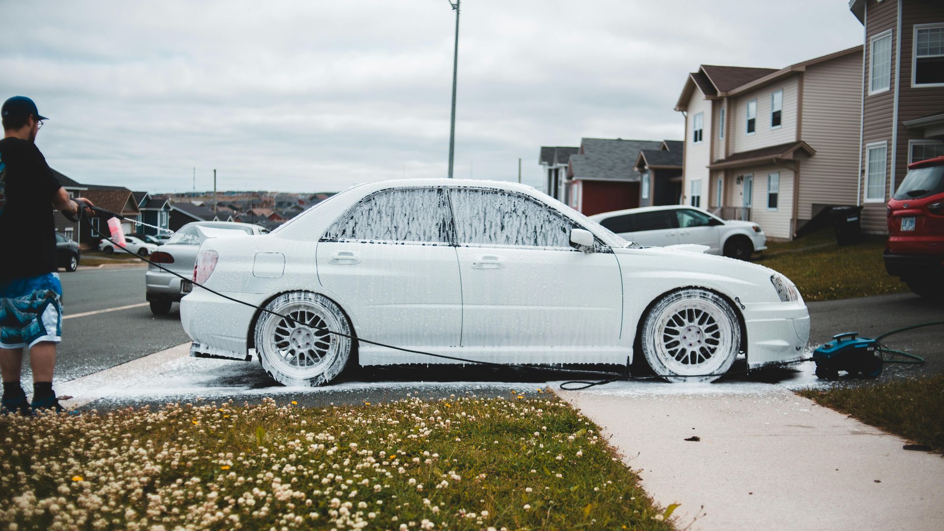 Foamed car on driveway and sidewalk being washed by male driver at daytime