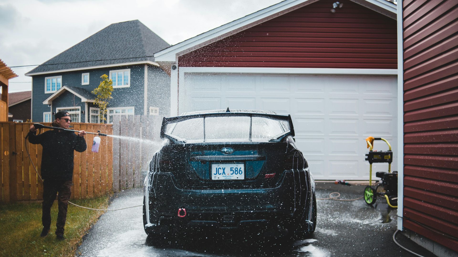 A man uses a hose to wash a sports car in a residential driveway, creating a splash effect.