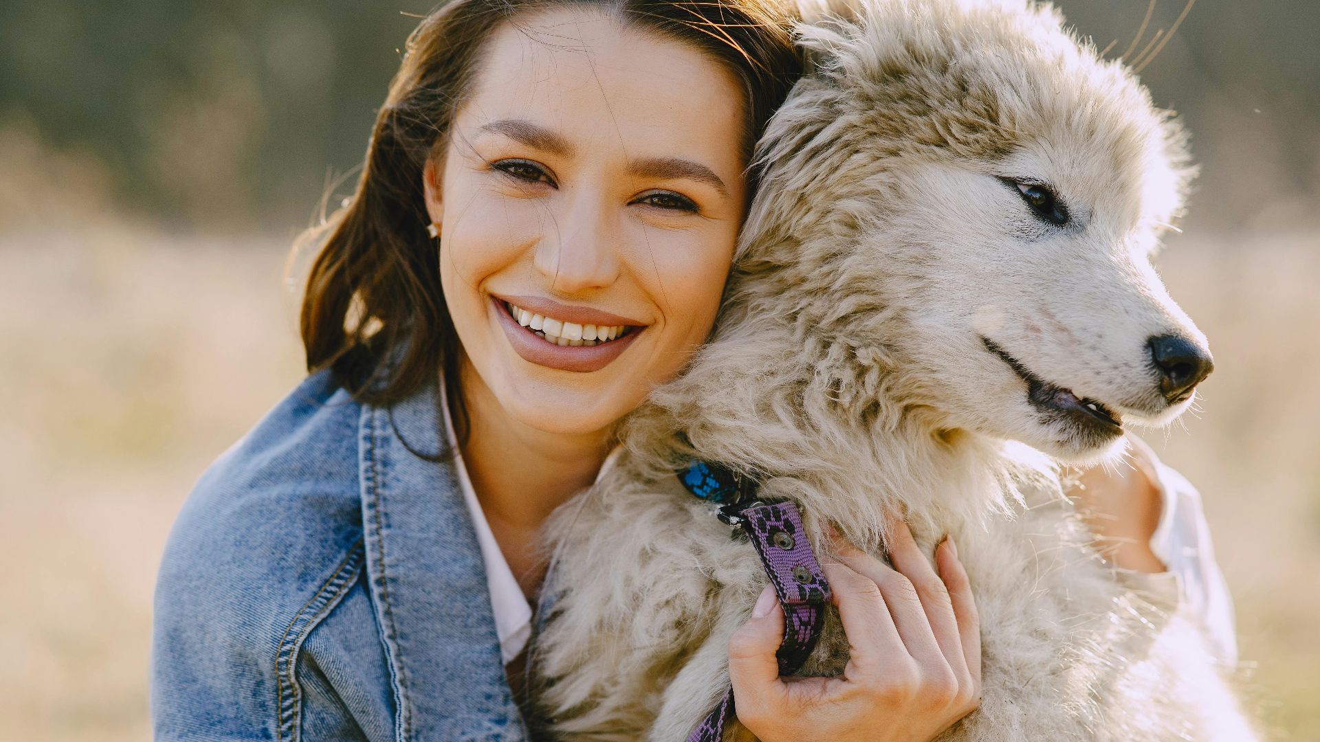 Joyful woman in a denim jacket hugging a fluffy dog outdoors, showcasing friendship and happiness.