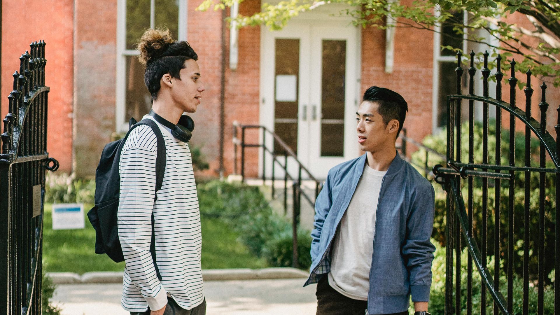 Full length positive young multiracial friends in casual clothes with backpacks talking and looking at each other while standing on street outside brick building