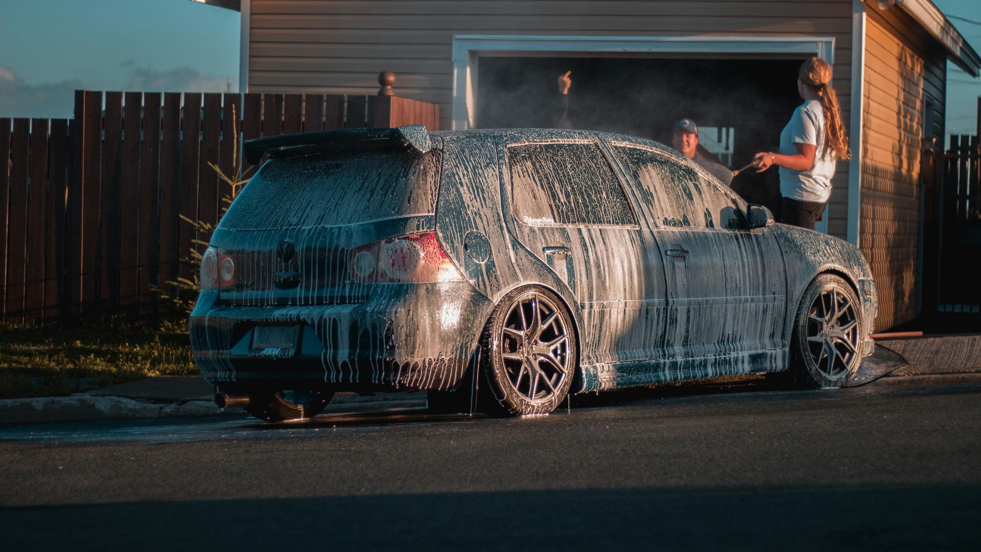 A family washes a car with soap suds on a suburban driveway during the day.