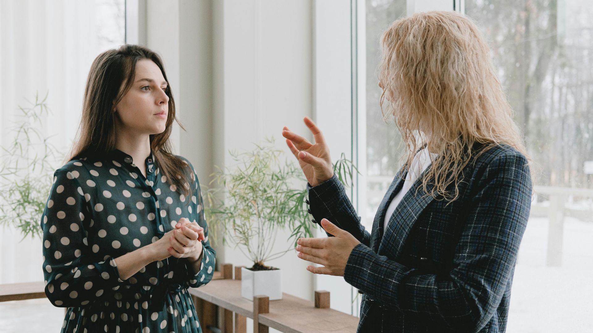 Two women engaging in a professional indoor conversation by a large window.