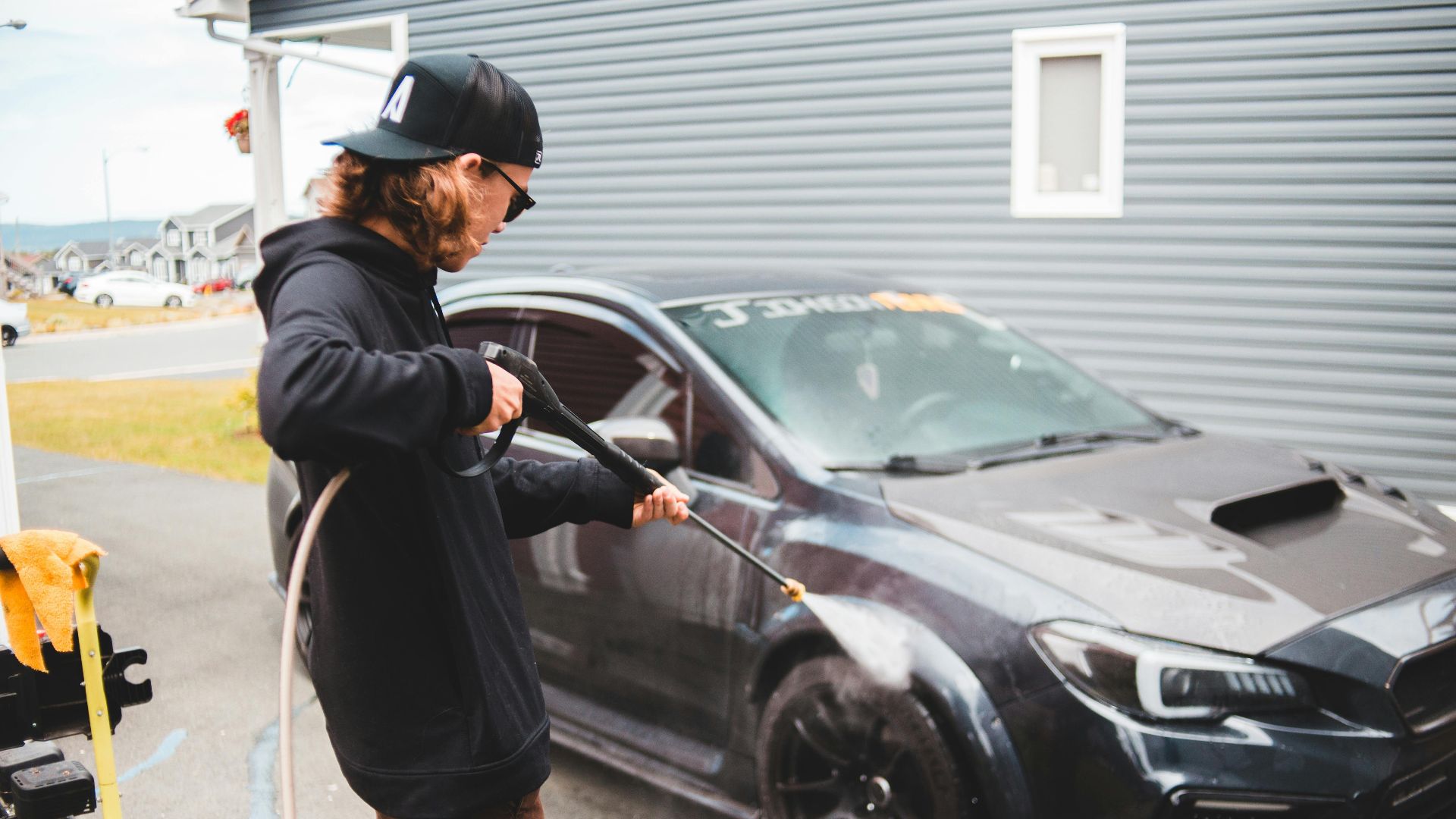 A man power washes a black sports car in a residential area, highlighting outdoor maintenance.