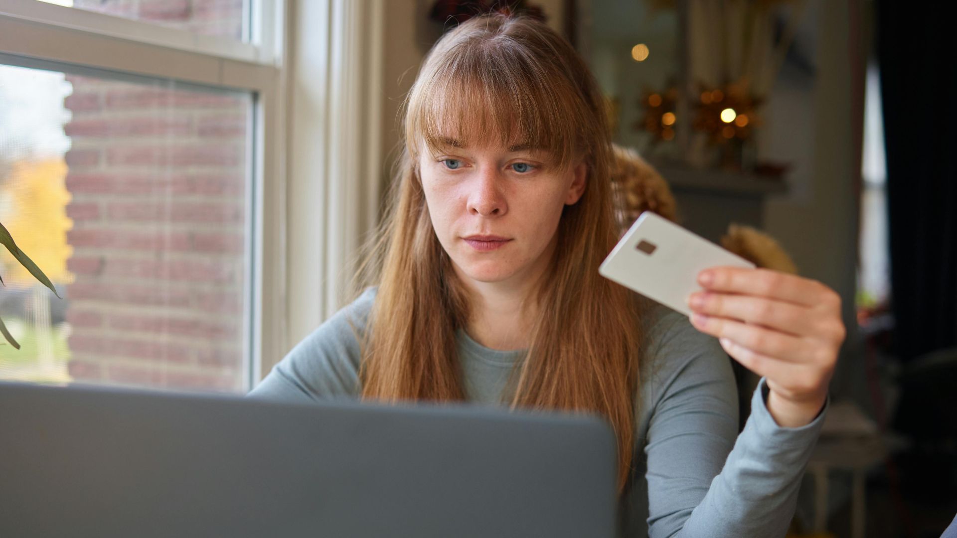 A young woman holds a credit card while using her laptop indoors.
