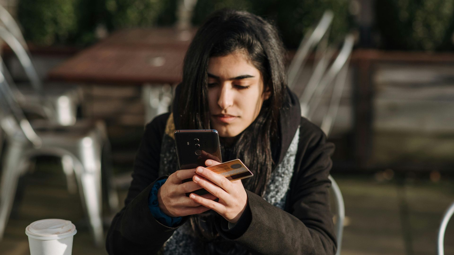 Young ethnic female purchaser with credit card shopping online on mobile phone at table with takeaway hot drink in cafeteria