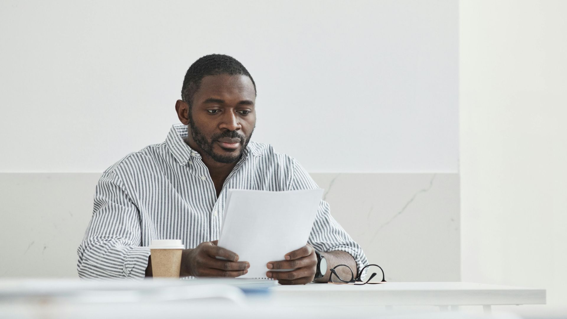 African American man sitting indoors, reading papers with a coffee cup nearby.