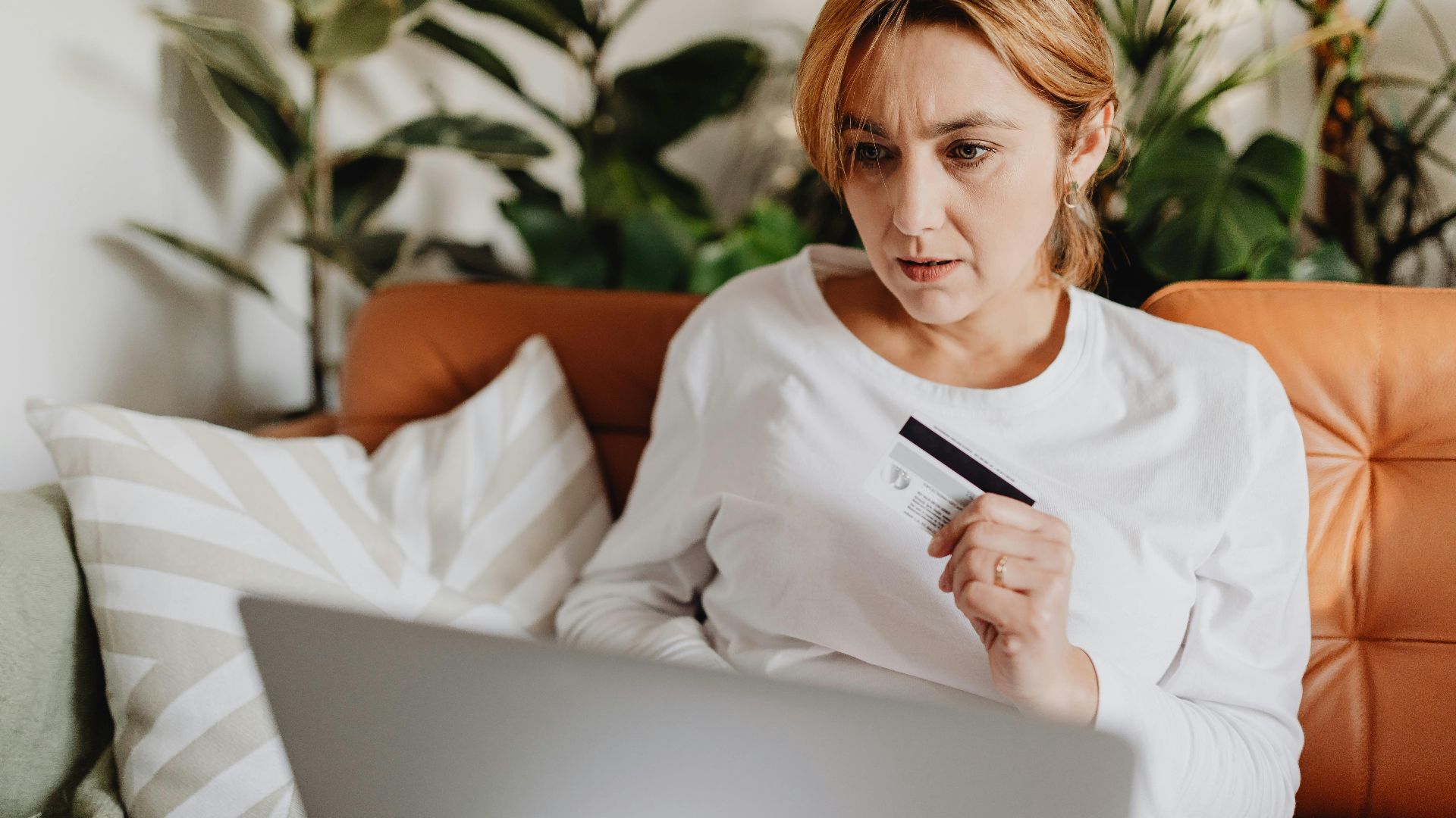 A woman sitting on a couch shopping online with a credit card and laptop in a cozy living room.