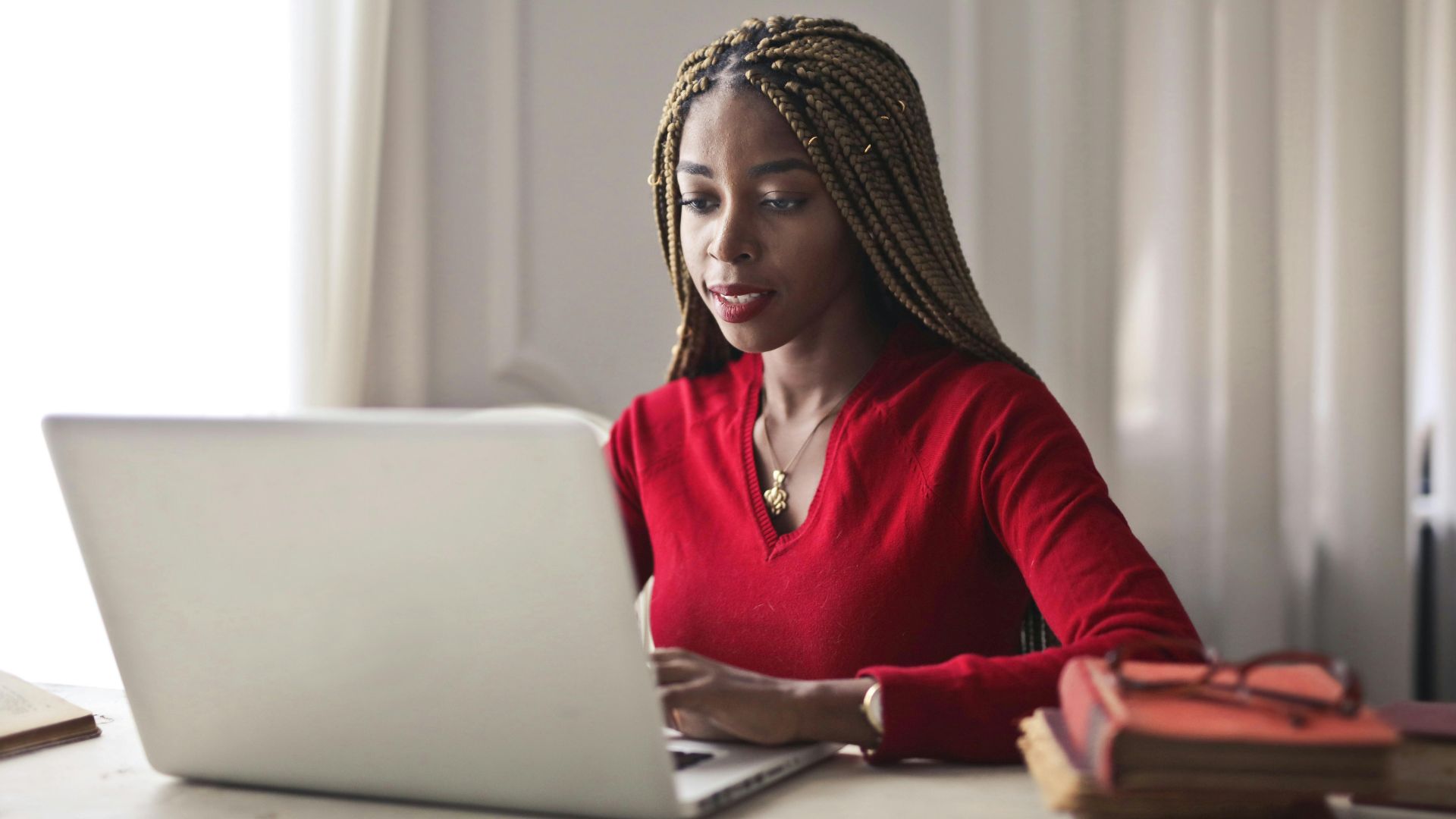 Confident woman in a red sweater working on a laptop in a bright, organized workspace.
