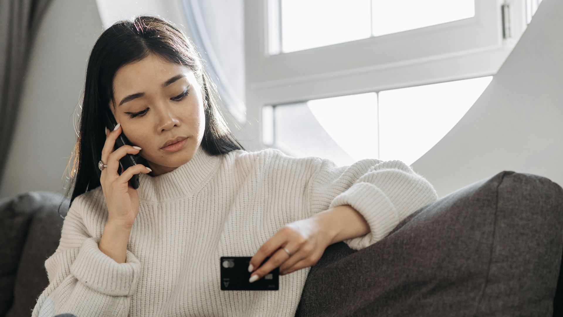 A woman sits on a sofa, multitasking with a phone and credit card, focusing on online shopping.