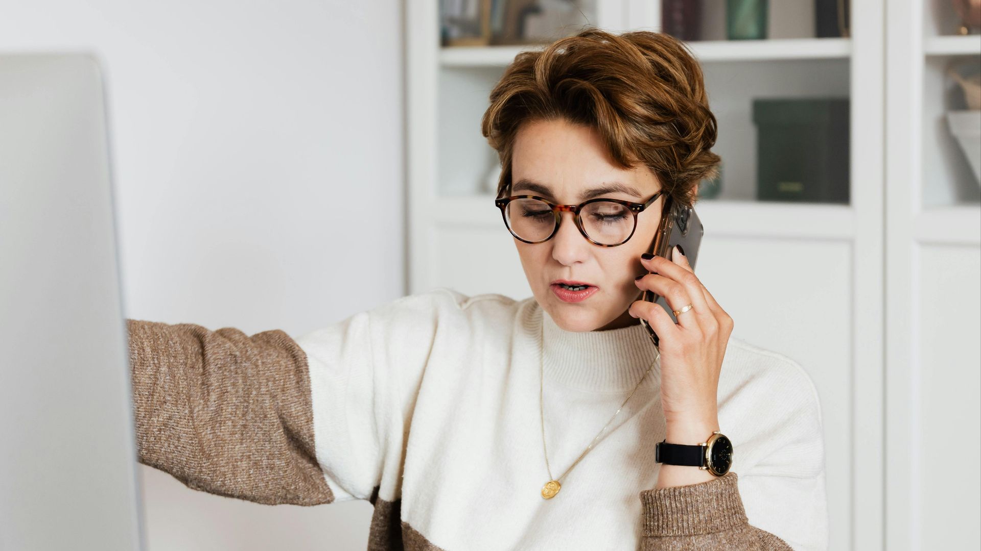 Female remote worker in eyeglasses talking on mobile with boss while working on project at home