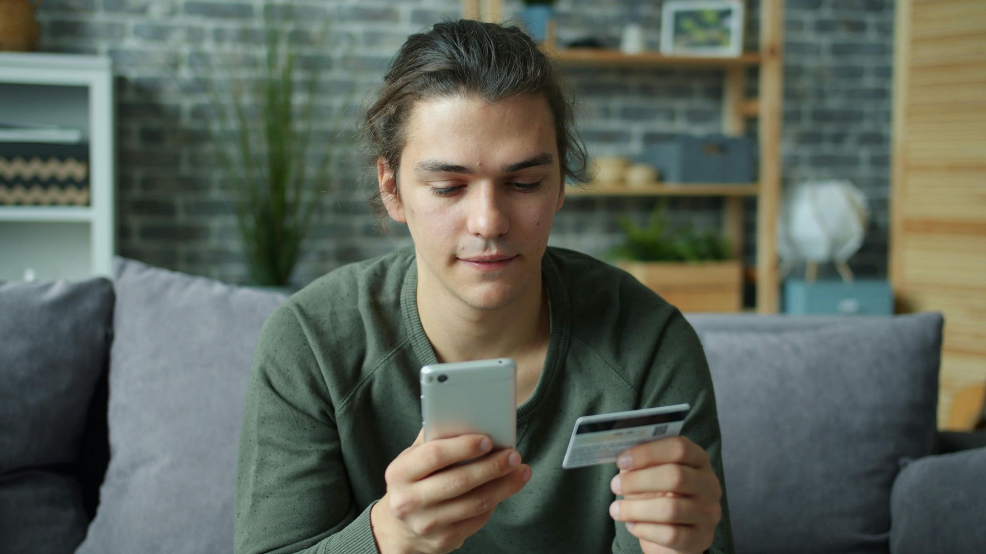 A young man using a smartphone and credit card for online shopping at home.