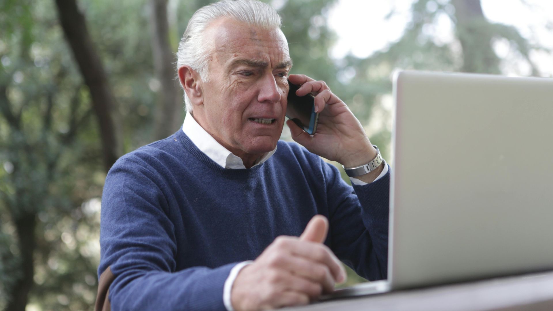 Elderly man using laptop and smartphone outdoors, showcasing modern connectivity.