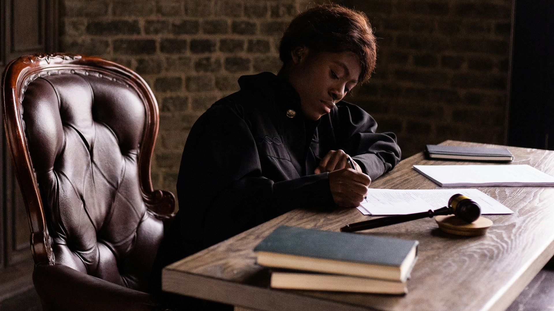 Black female judge in judicial robe writing at desk in an office setting, focusing on legal work.