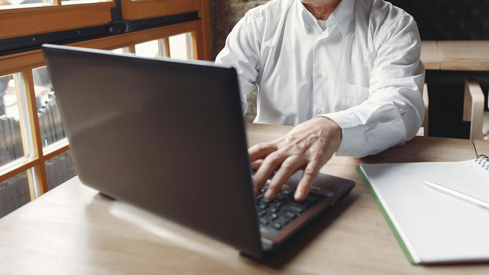 A senior man focuses on his laptop at a wooden table in a bright, cozy cafe during the day.