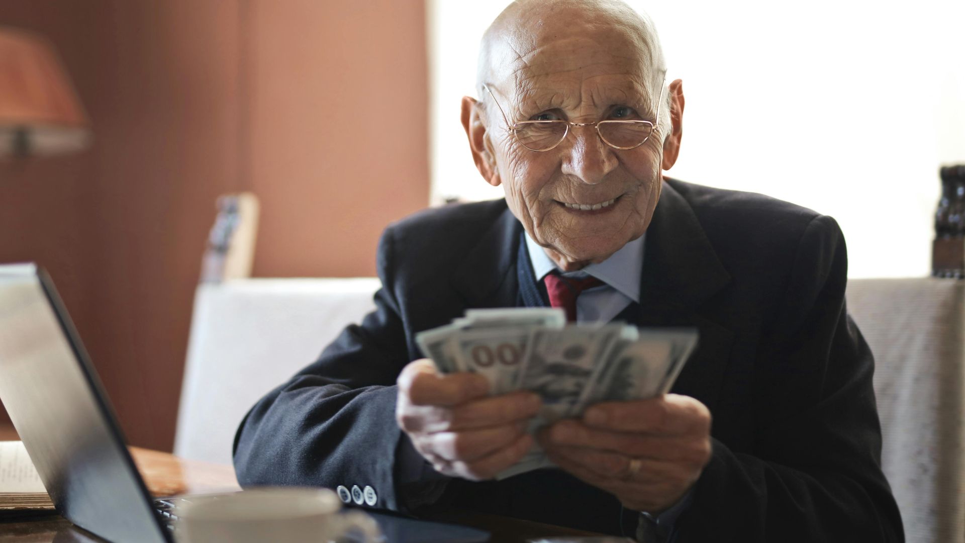 Elderly man smiling while counting cash at a home office setting.