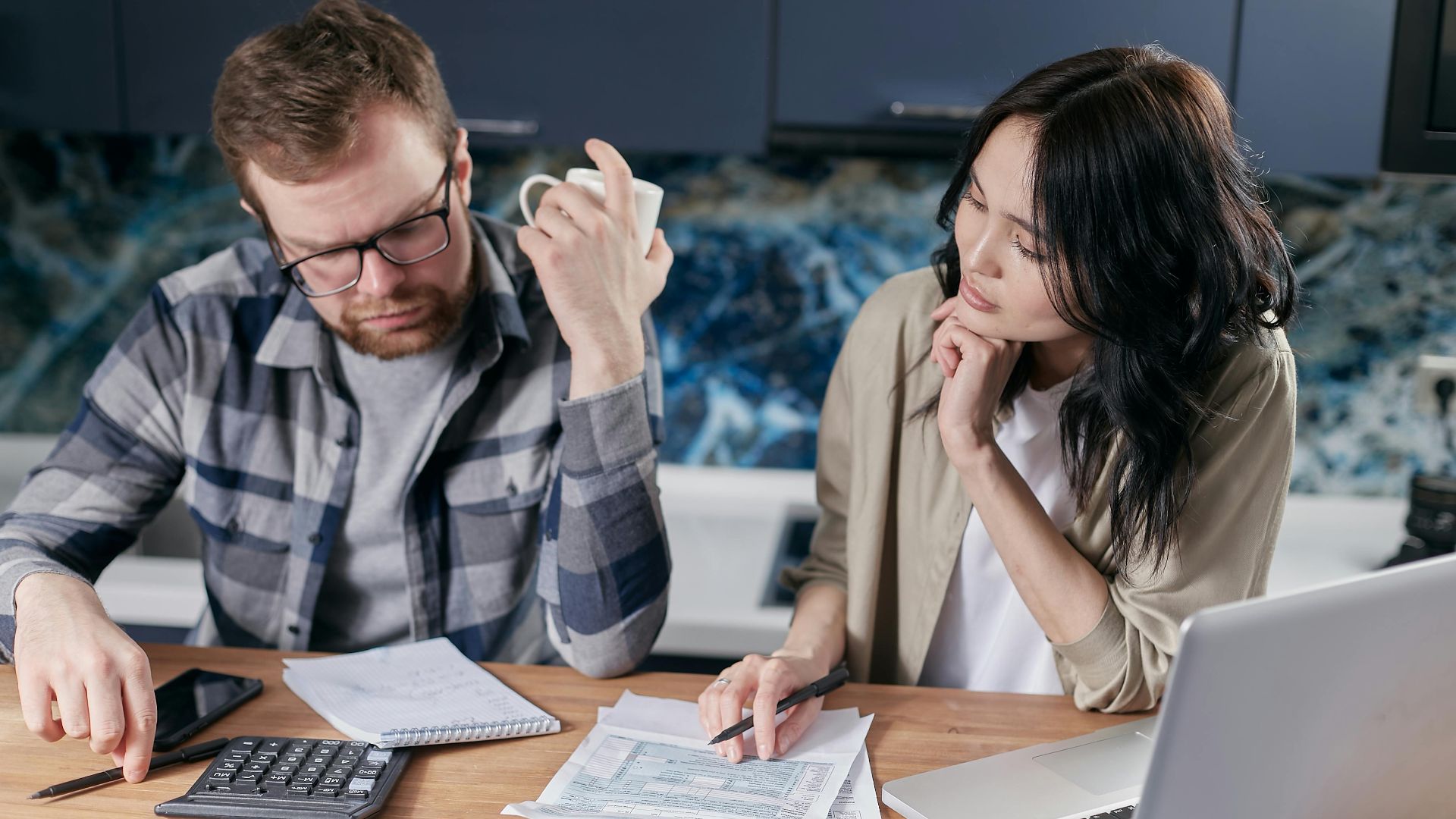 A couple reviews bills and documents on a laptop while discussing household finances.