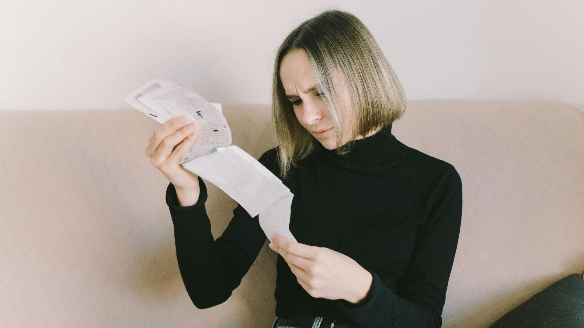 Concerned woman in black sweater examining bills on beige sofa indoors.