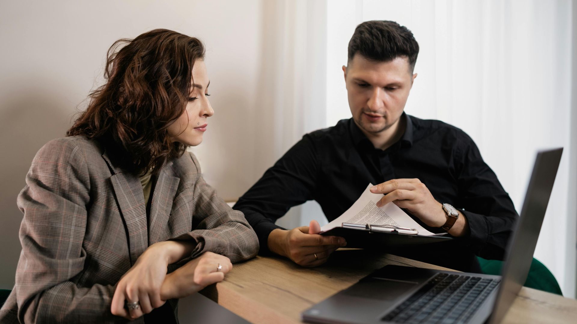 Two professionals discussing documents at a desk with a laptop, focused on collaboration.