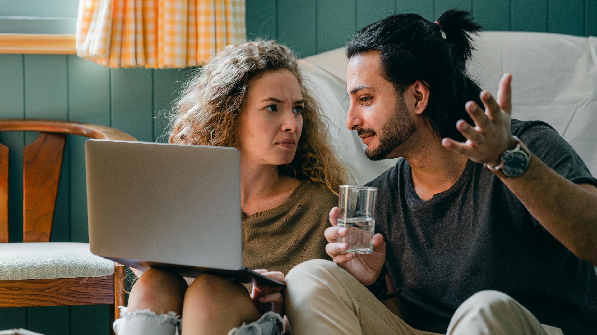 A couple sits on the floor, engaged in conversation, using a laptop in a cozy apartment.