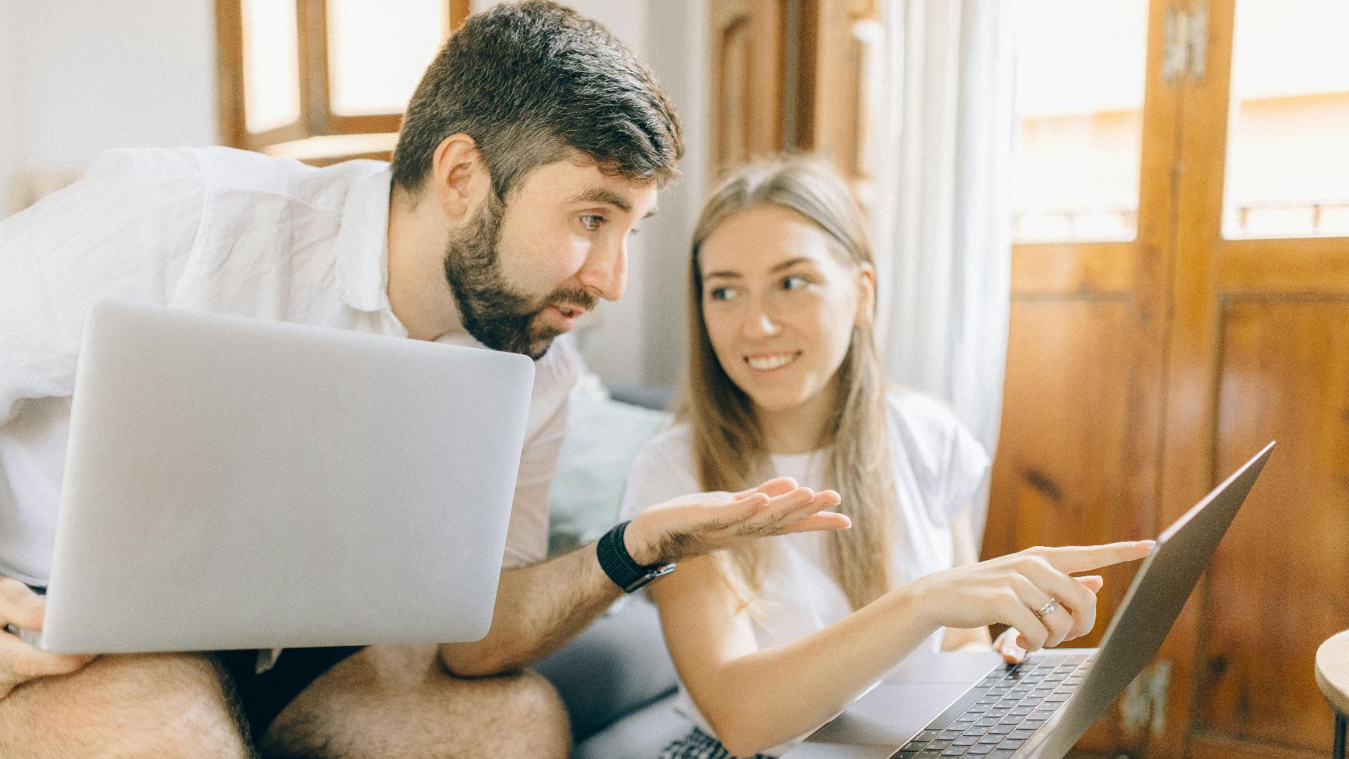 Young couple sitting together, discussing something on their laptops in a cozy room.