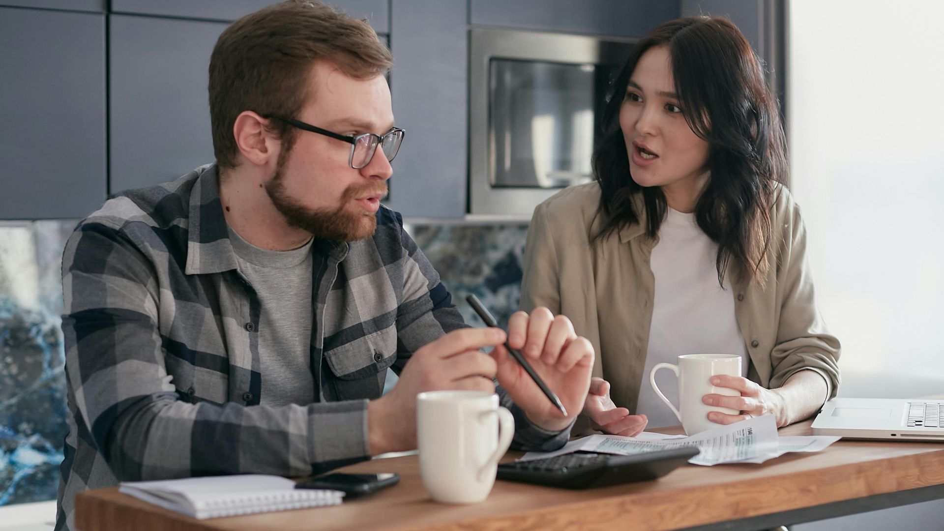 A couple managing finances with gadgets, documents, and coffee mugs at home.