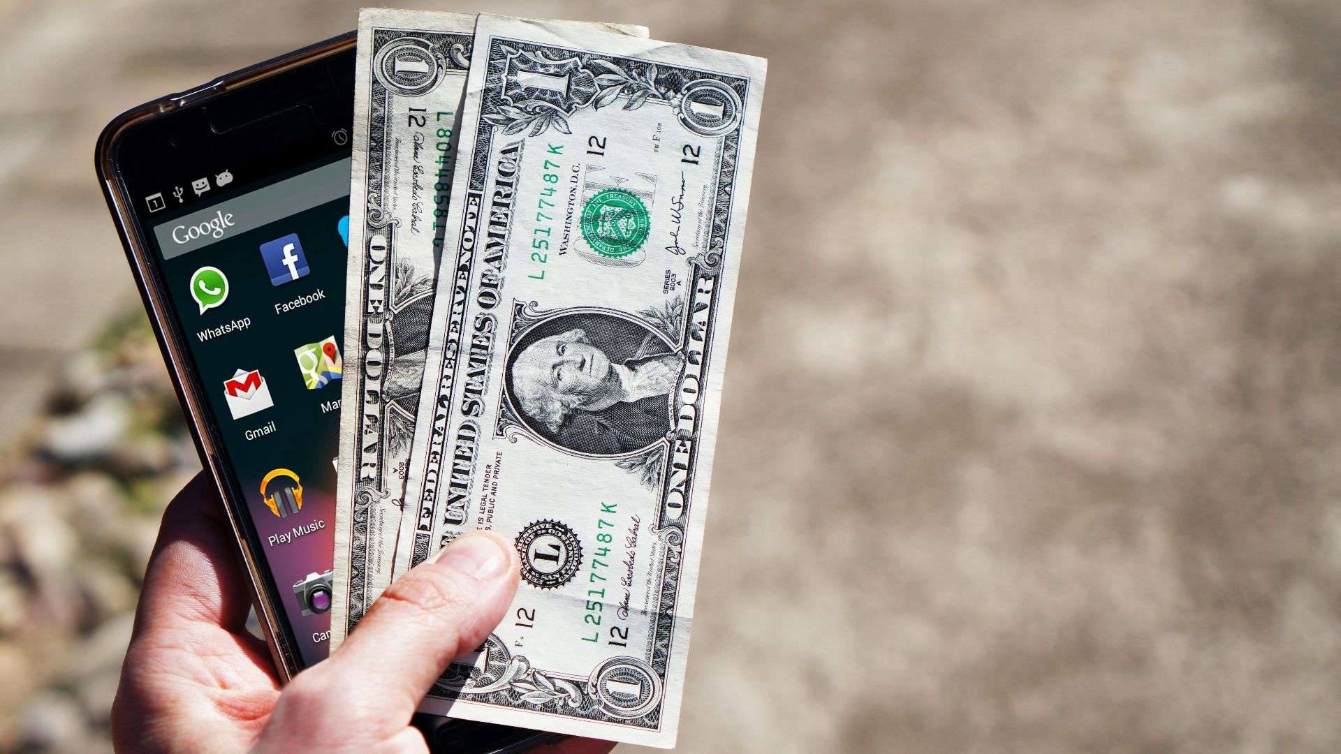 Close-up of a hand holding US dollar bills and a smartphone outdoors, showcasing financial technology.