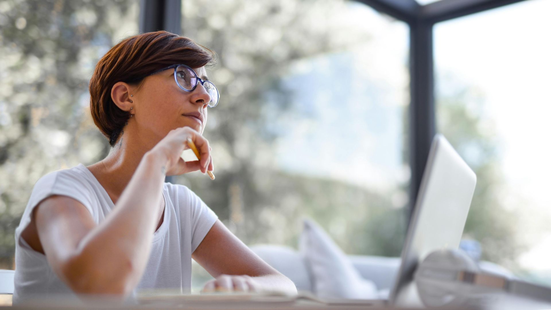 Thoughtful woman with glasses and short hair looking out window in a modern room.