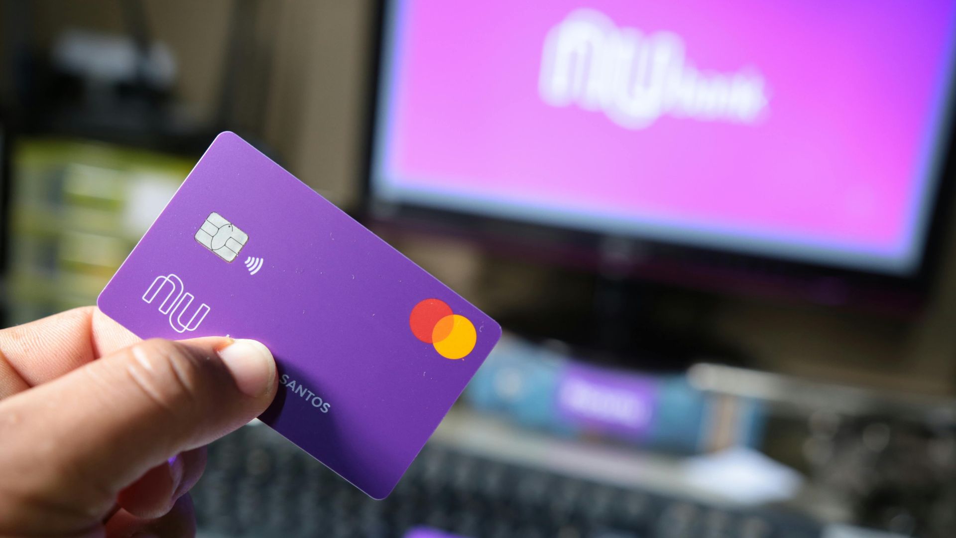 A close-up of a hand holding a purple bank card in front of a computer screen.
