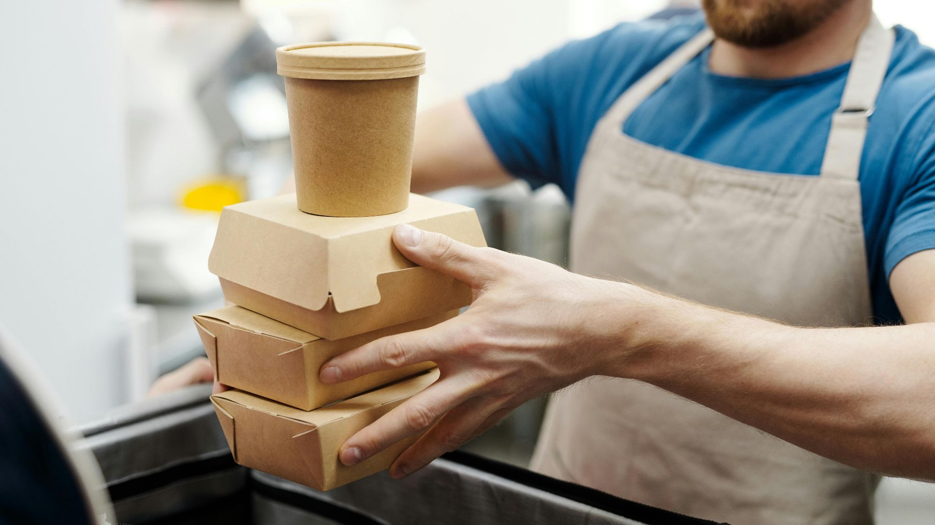 Man wearing apron stacks eco-friendly food containers for takeaway delivery in a kitchen setting.