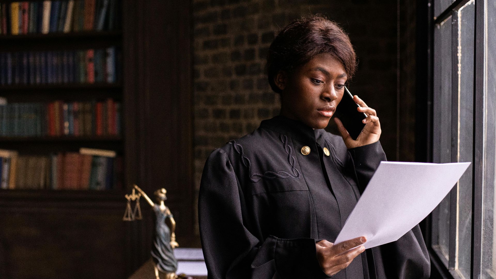 African American judge in robe, reading document and talking on phone by the office window.