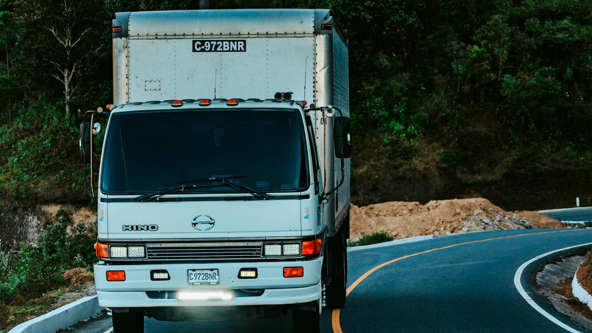 A white truck driving on a winding road surrounded by dense green forest plants.