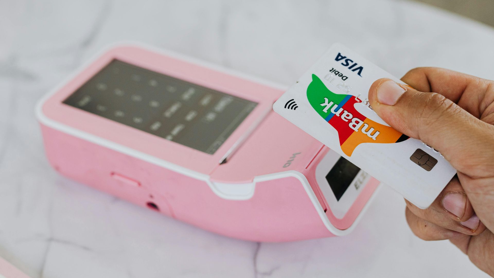 A hand using a Visa card for a contactless payment on a pink terminal.