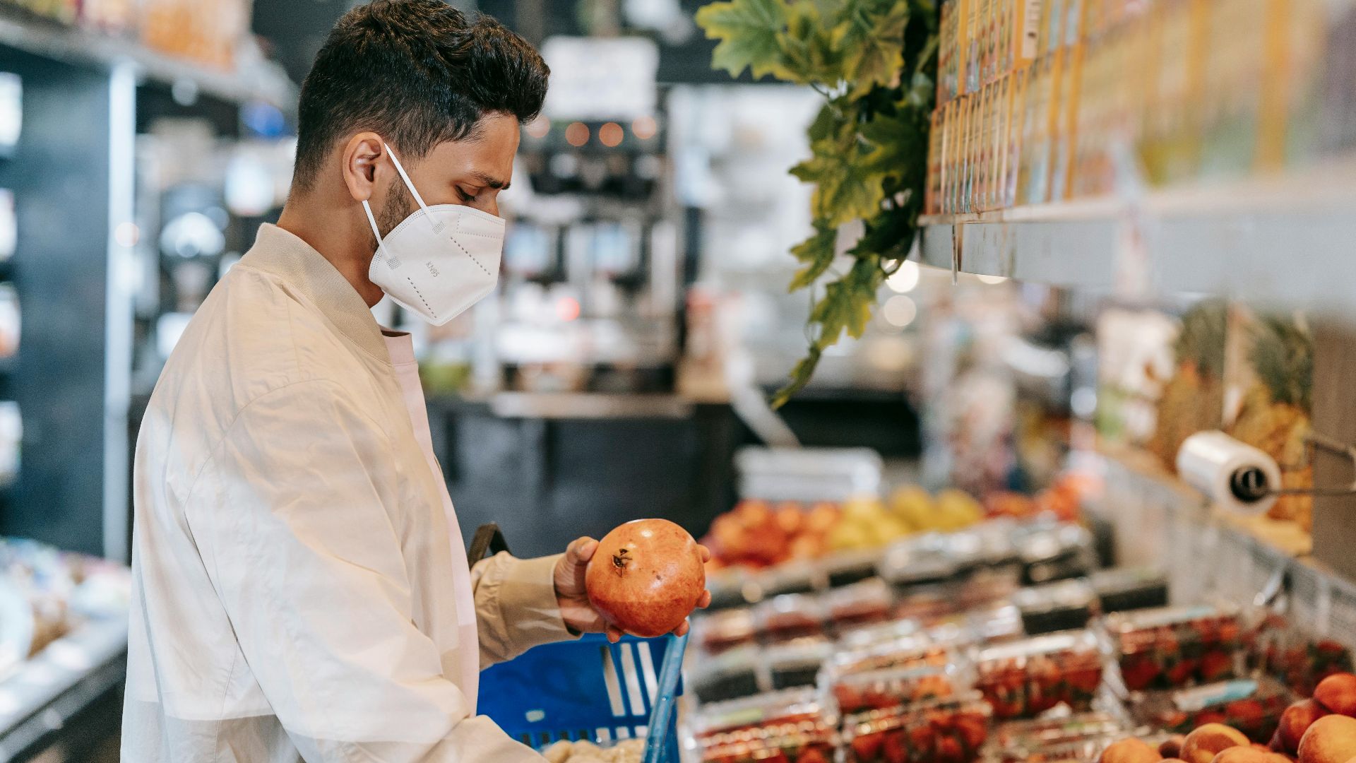 Side view of young Hispanic man in protective mask choosing fresh pomegranate from stall in supermarket during coronavirus pandemic