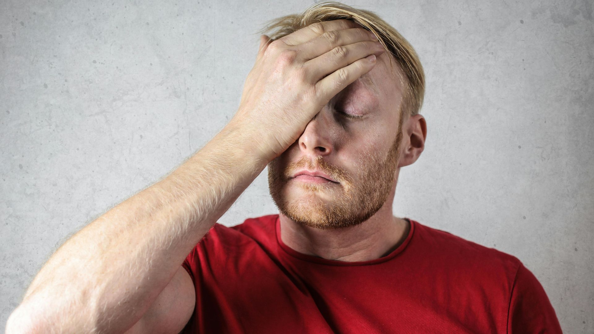 A frustrated man in a red shirt holds his head in stress against a neutral background.