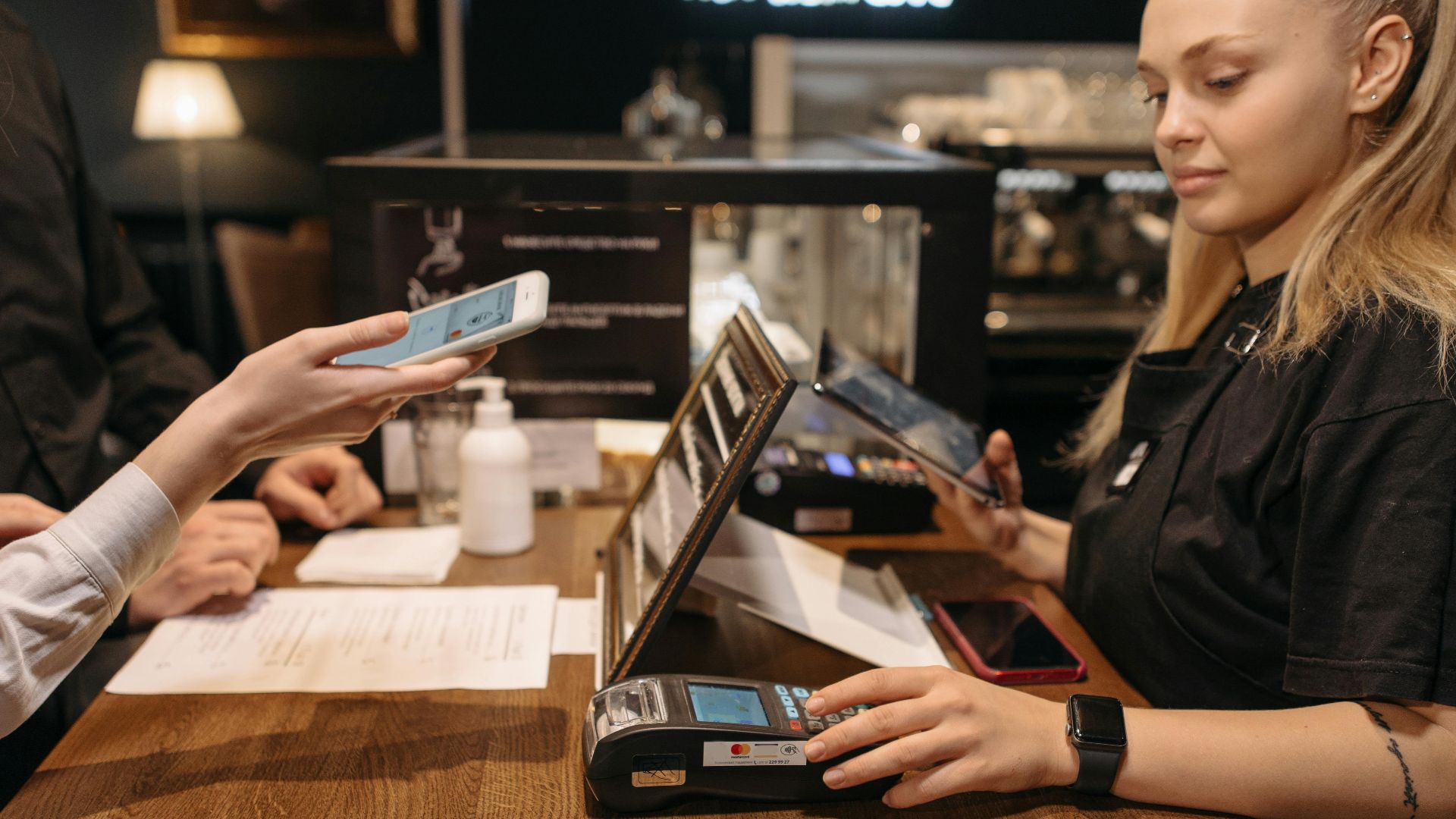 Customer using mobile phone for contactless payment at a coffee shop counter with a barista.