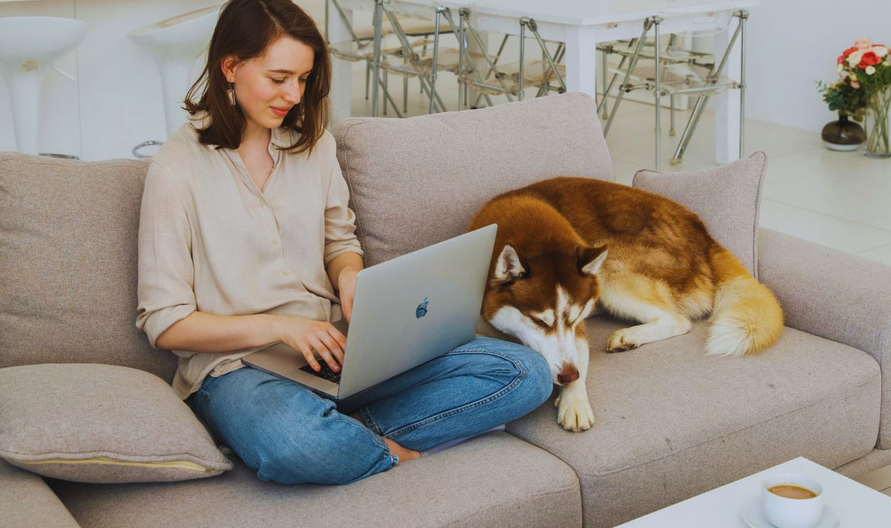 Woman Sitting on Gray Couch Using Laptop Beside A Dog
