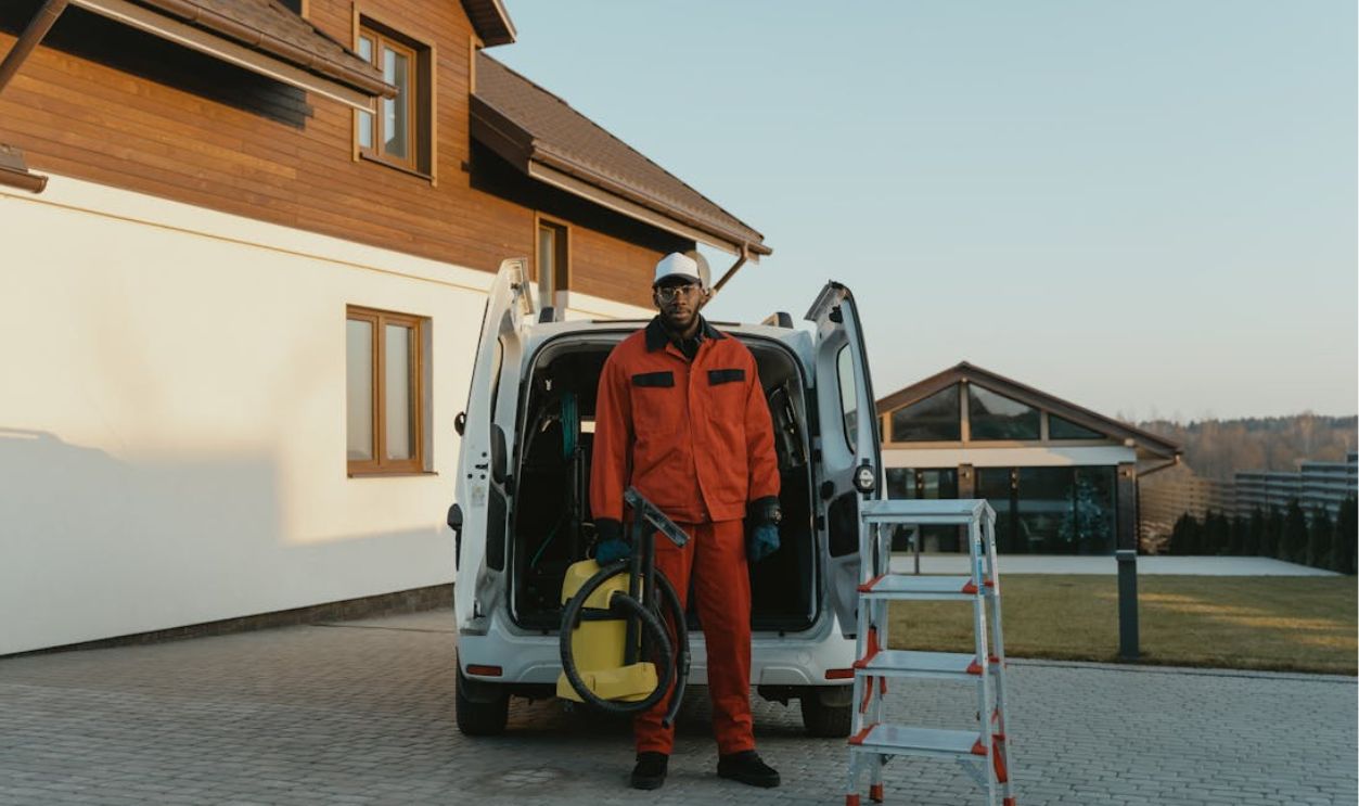 Man in Working Clothes With A Vaccum Cleaner Standing Beside A Service Van and Stepladder
